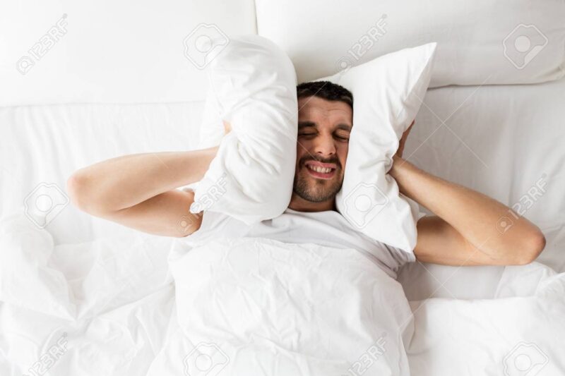 Homem na cama, tapando os ouvidos com os travesseiros enquanto ouve ruídos